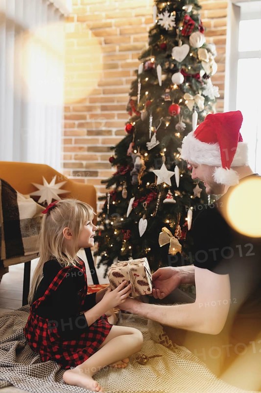 Young father in red Santa hat giving Christmas gift to his smiling daughter near decorated Christmas tree. Girl dressed in festive red-black Christmas outfit. They smiling, happy because of New Year