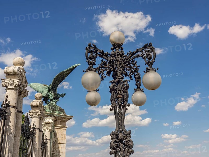 Wrought iron street lamp with ball lamps in front of a blue sky and a winged statue, budapest, danube, hungary