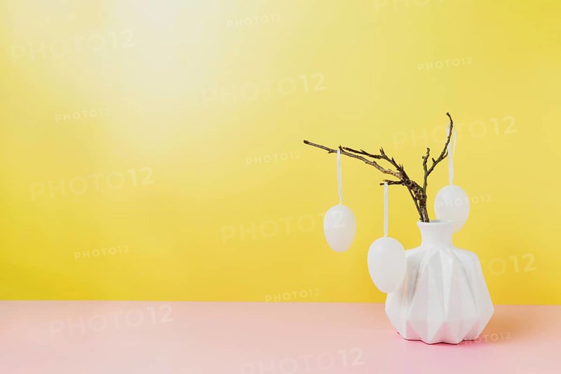 Happy Easter holiday, coming spring concept. Tree branches, flower twigs in white vase or jar with hanging eggs. Yellow and pink table background with copy space. Easter preparation, greeting card