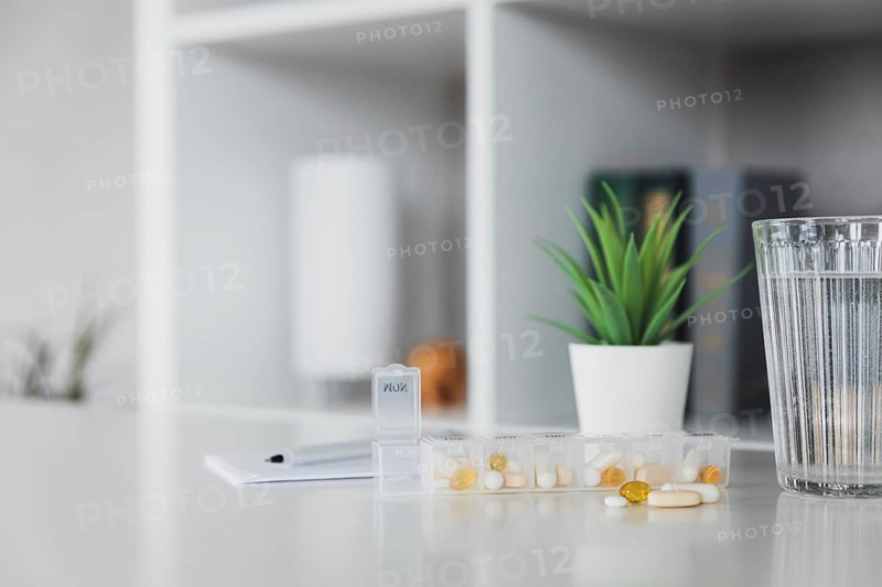 Closeup of medical pill box with doses of tablets for daily take a medicine for treatment, cure the disease. Glass cup of water on table, gray background. Green flowerpot or plant. Pen and notepad