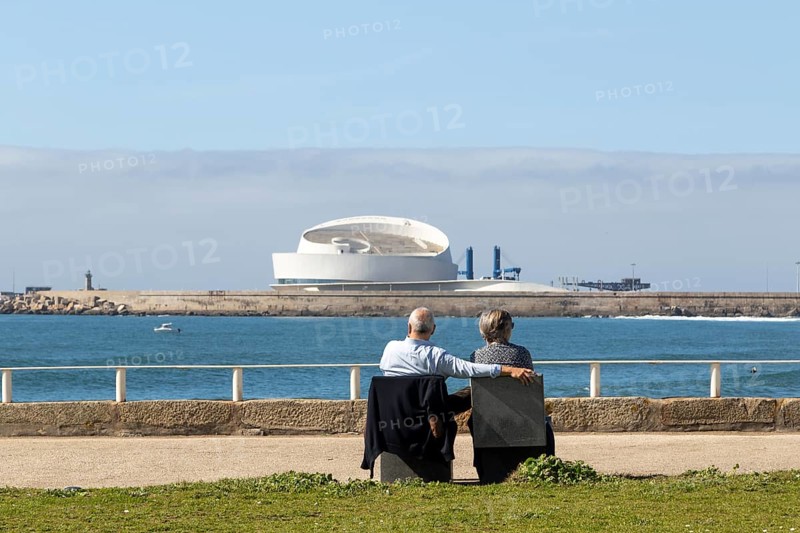 A couple of senior citizens sitting on a park bench looking at the Porto Leixões Cruise Terminal on the seafront promenade at Praia de Matosinhos beach in Matosinhos, Norte region, Porto district, Portugal, Europe