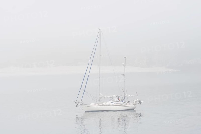 Sailboat, sailing boat, yacht with lowered sails anchored near beach during bad visibility due to thick fog, dense mist