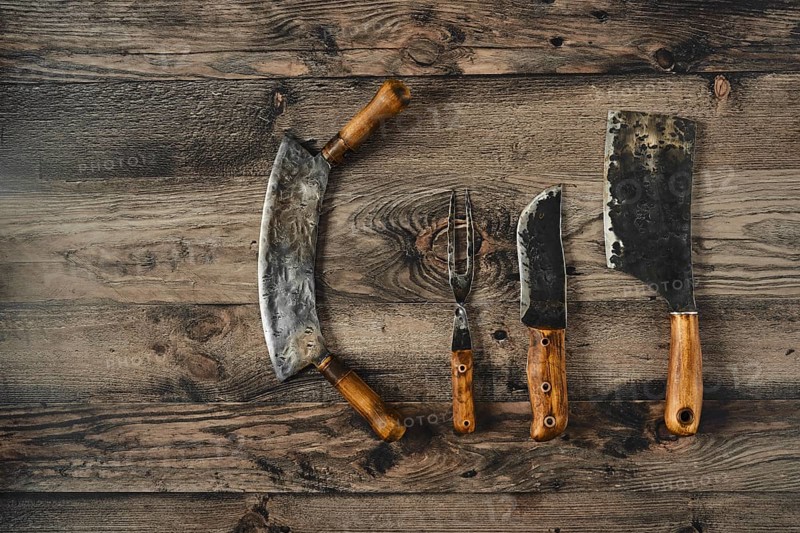 A collection of vintage kitchen knives, including a cleaver and a carving knife, is displayed on a weathered wooden table. The tools showcase worn wooden handles and unique blade designs