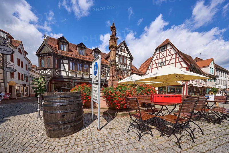 Market fountain with geraniums, tables, chairs and parasols of a restaurant and half-timbered houses under a blue sky with cumulus clouds on the market square in Michelstadt, Odenwald, Odenwaldkreis, Hesse, Germany, Europe