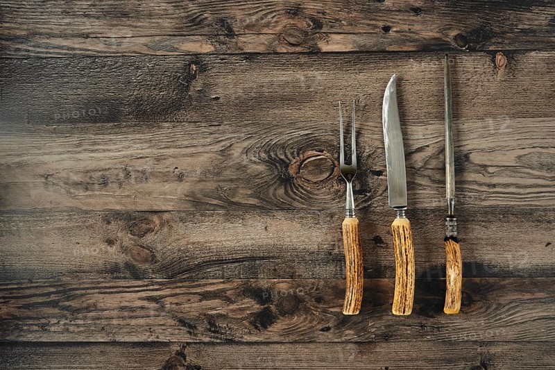 Three essential barbecue tools on a rustic kitchen table made of dark wood, knife, fork and sharpener. The setting suggests a cozy cooking environment