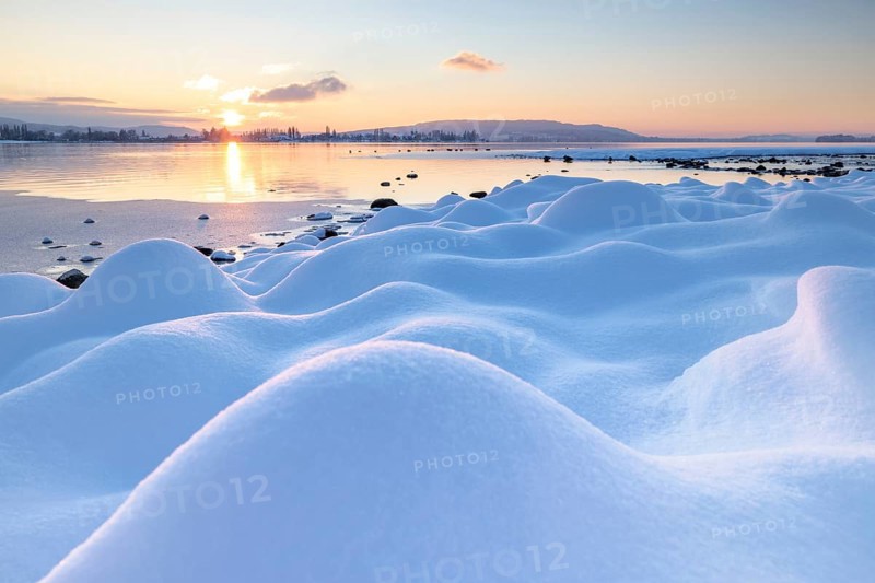 Gentle hills of snow stretch towards the lake, where the sun sets over the mountains, Seegarten, Allensbach, Lake Constance, Baden-Württemberg, Germany, Europe