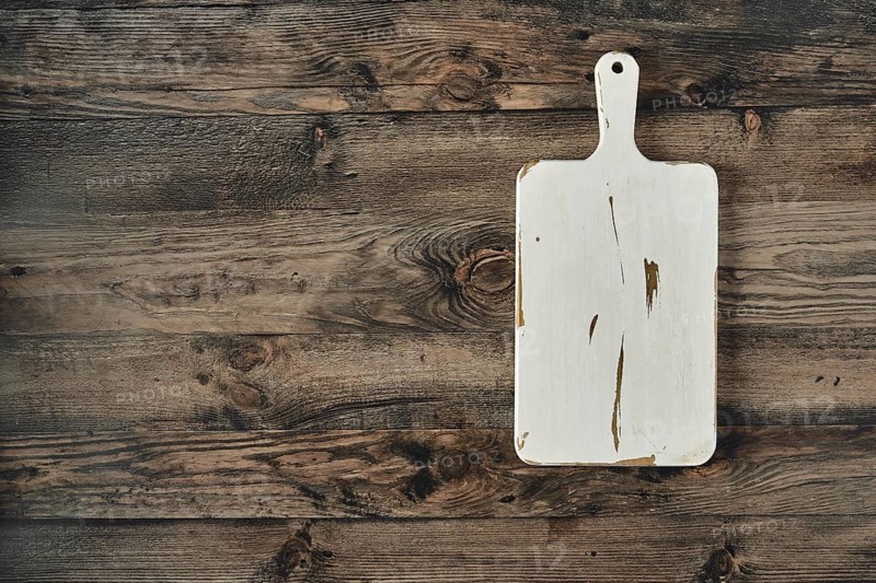 A well-worn wooden cutting board rests on a dark, rustic wooden table. The board shows signs of use with visible marks and scratches, adding character to a cozy kitchen environment