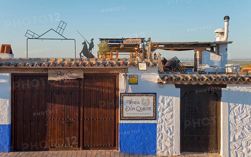 Rustic building with signs and white-blue walls, windmills and sun in the background under a clear sky, Campo de Criptana, Ciudad Real province, Castilla-La Mancha, Don Quixote Route, Spain, Europe
