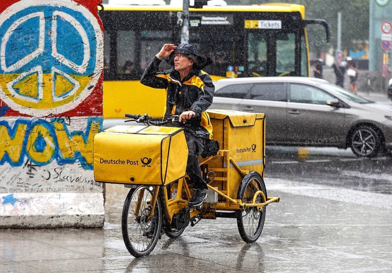A DHL mail carrier rides his e-bike along a pavement in heavy rain, Berlin, 23 06 2023