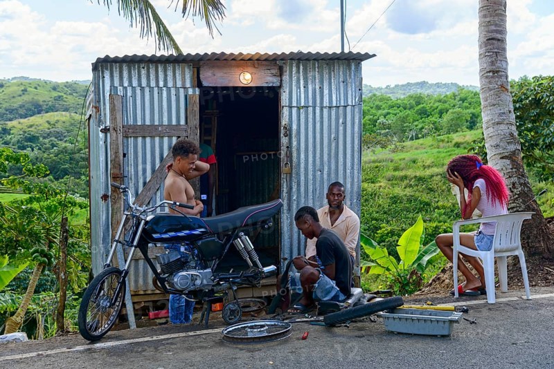 A group of people repairing a motorbike in front of a metal hut surrounded by green landscape, mending tyres, Typical workshop made of corrugated iron, Dominican Republic, Hispaniola, Caribbean, America, Central America