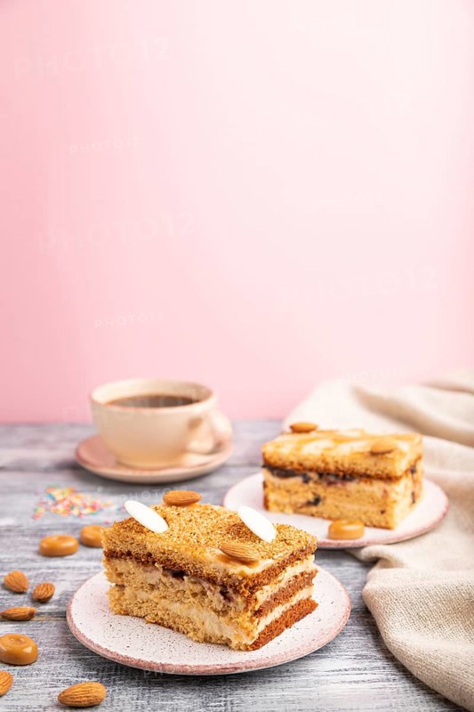 Honey cake with milk cream, caramel, almonds and a cup of coffee on a gray and pink background and linen textile. Side view, close up, selective focus, copy space