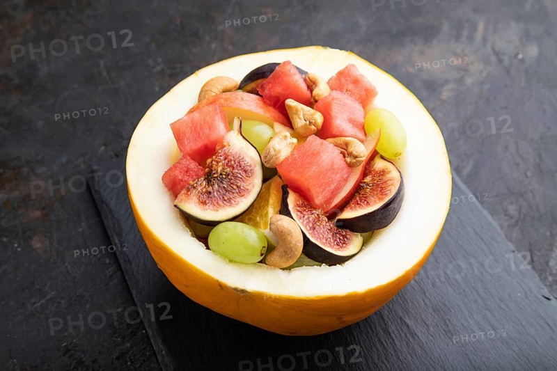 Vegetarian fruit salad of watermelon, grapes, figs, pear, orange, cashew on slate board on a black concrete background. Side view, close up