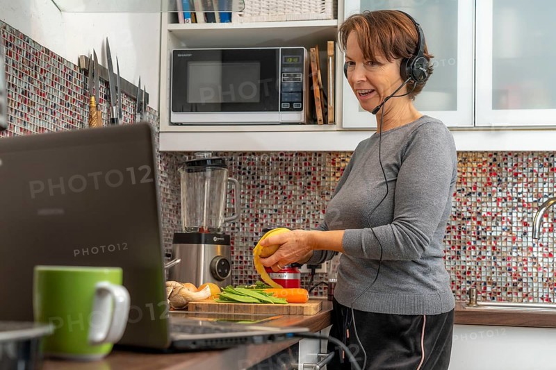 Woman, mid-50s, works from home, with laptop and communicates with colleagues via headset, home office, in the kitchen, while cooking