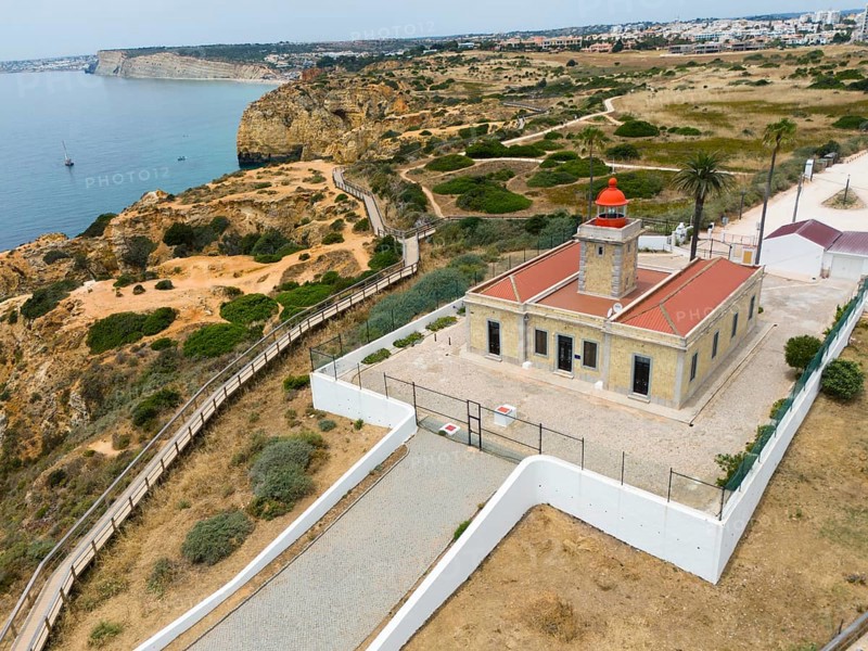 A lighthouse on the coast, surrounded by cliffs, the sea and town in the background. A path leads through the natural vegetation, aerial view, Ponta da Piedade, point of mercy, Lagos, Algarve, Portugal, Europe