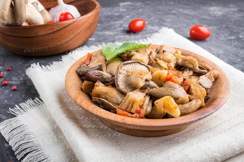 Fried oyster mushrooms with tomatoes in wooden plate on black concrete background. side view, close up