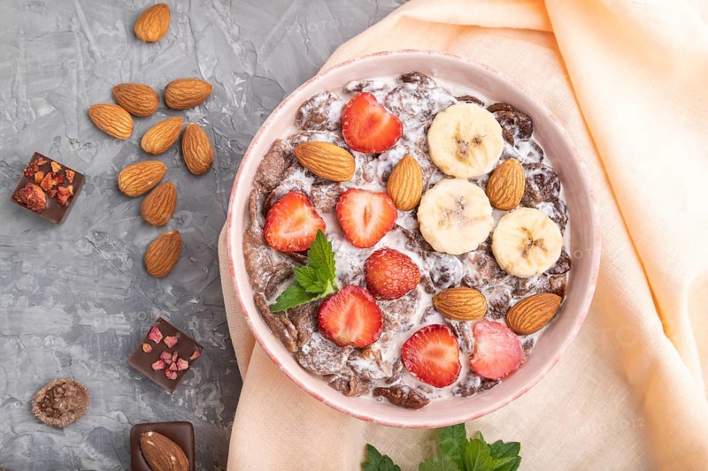Chocolate cornflakes with milk, strawberry and almonds in ceramic bowl on gray concrete background and orange linen textile. Top view, flat lay, close up