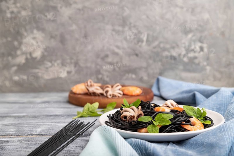 Black cuttlefish ink pasta with shrimps or prawns and small octopuses on gray wooden background and blue textile. Side view, copy space, selective focus