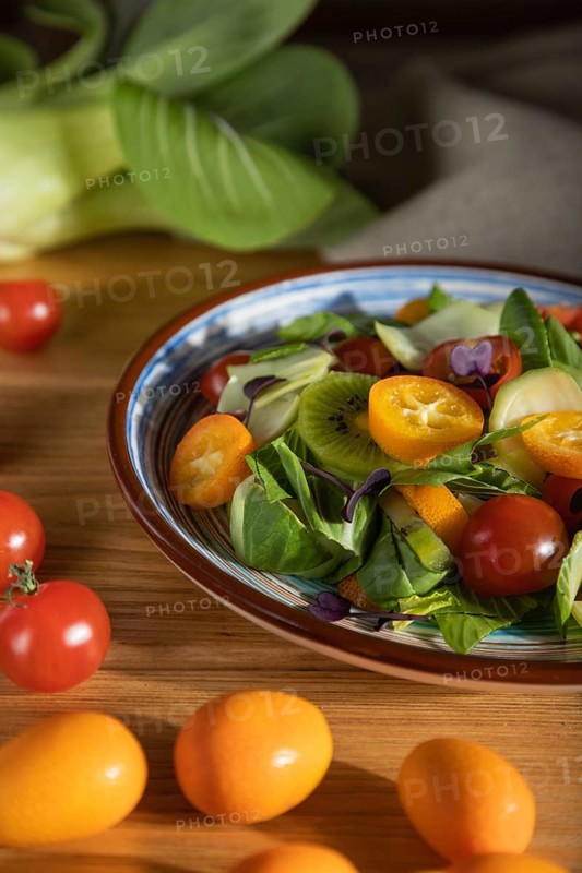 Vegetarian salad of pac choi cabbage, kiwi, tomatoes, kumquat, microgreen sprouts on a wooden background and linen textile. Hard light, contrast. Side view, close up, selective focus