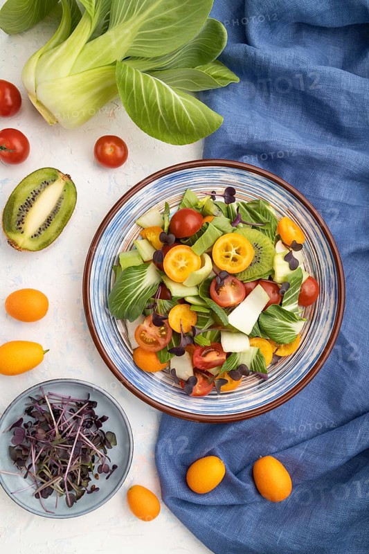 Vegetarian salad of pac choi cabbage, kiwi, tomatoes, kumquat, microgreen sprouts on a white concrete background and blue linen textile. Top view, flat lay, close up