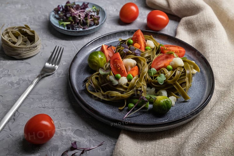 Tagliatelle green spinach pasta with tomato, pea and microgreen sprouts on a gray concrete background and linen textile. Side view, close up, hard light