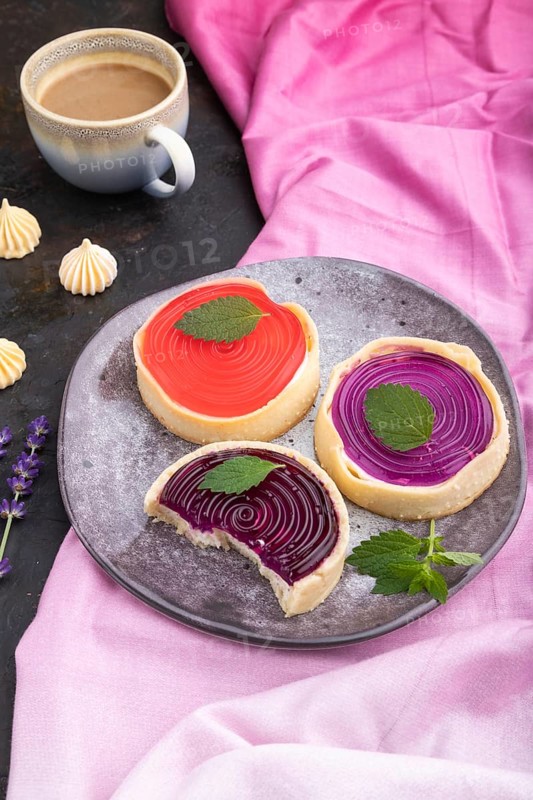 Sweet tartlets with jelly and milk cream with cup of coffee on a black concrete background and pink textile. Side view, close up