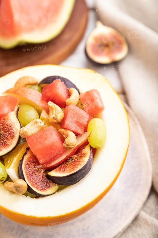 Vegetarian fruit salad of watermelon, grapes, figs, pear, orange, cashew on white wooden background and linen textile. Side view, close up, selective focus