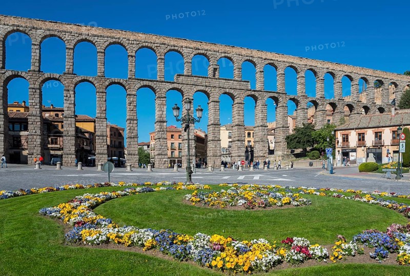 A historic stone aqueduct with arches spans a city under a bright blue sky next to a colourful flowerbed, Aqueduct, Segovia, Castilla y León, Leon, Spain, Europe