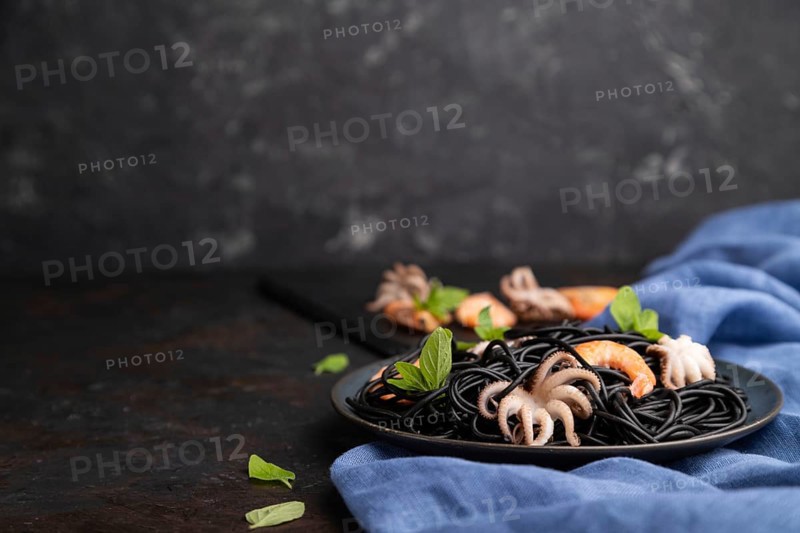 Black cuttlefish ink pasta with shrimps or prawns and small octopuses on black concrete background and blue textile. Side view, copy space, selective focus