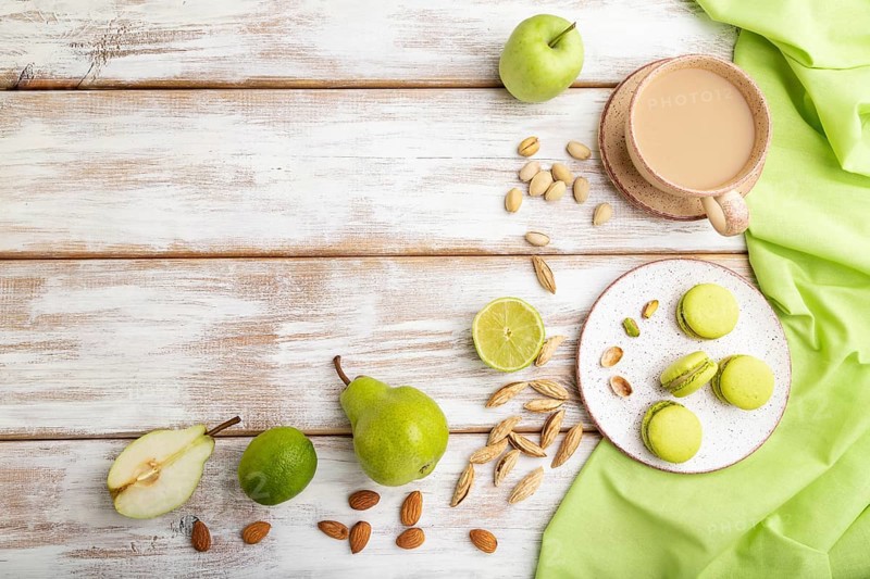 Green macarons or macaroons cakes with cup of coffee on a white wooden background and green linen textile. Top view, flat lay, copy space