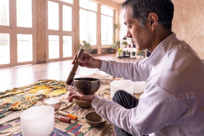 A middle-aged Asian man sits in profile, concentrating on gently vibrating a brass bowl in a softly lit room. Ancient practice of sound meditation in a quiet atmosphere