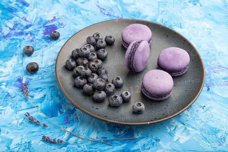 Purple macarons or macaroons cakes with blueberries on ceramic plate on a blue concrete background. Side view, close up