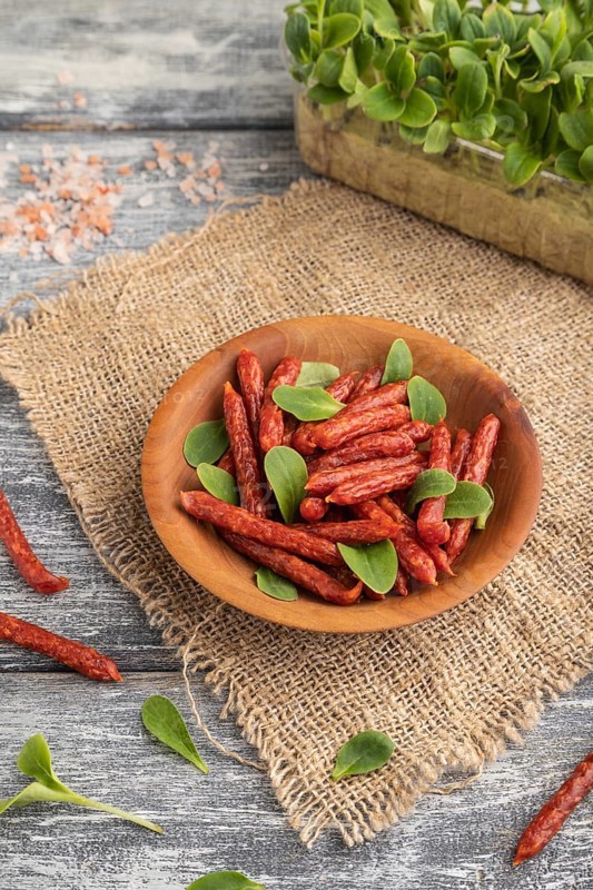 Small smoked sausage with borage microgreen, salt and pepper on gray wooden background and linen textile. Side view, close up