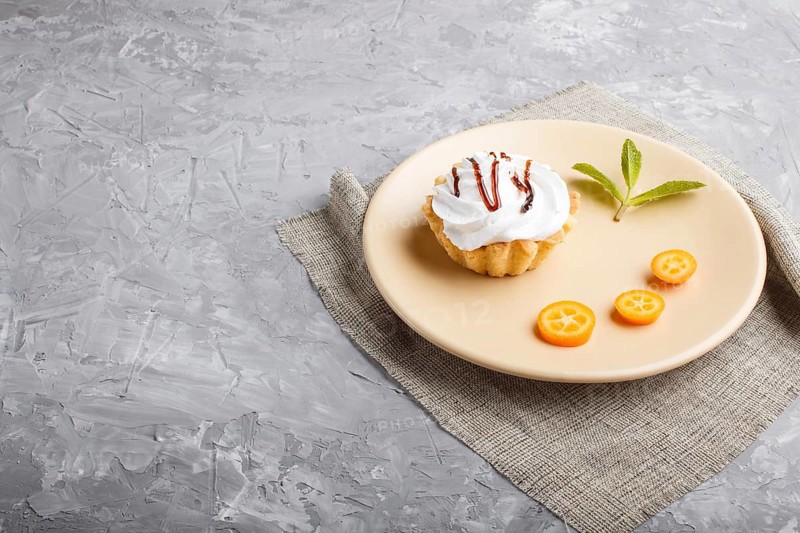 Cake with whipped egg cream on a light brown plate with kumquat slices and mint leaves on a gray concrete background with linen napkin copy space, side view