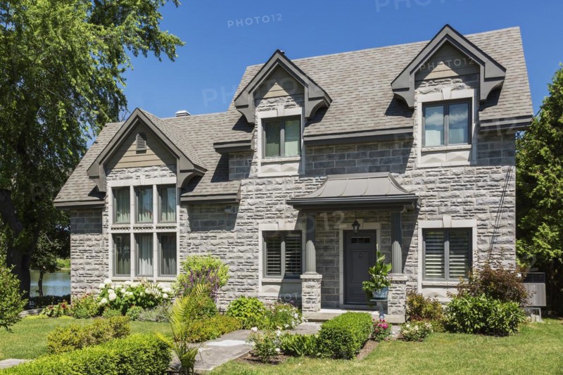 Contemporary tan brick house facade with brown trim and nuanced asphalt shingles roof and landscaped front yard in summer, Quebec, Canada, North America
