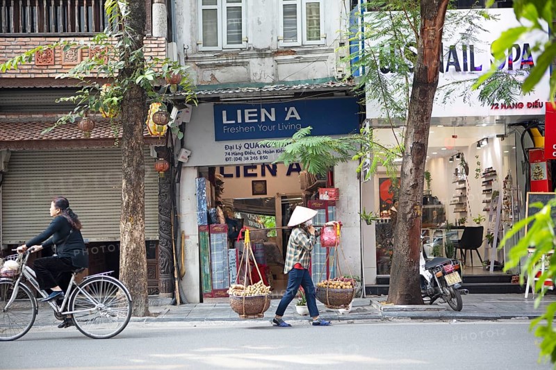 Vietnamese saleswoman in a conical hat shoulders cassava on a carrying pole through the Old Quarter of Hanoi, Vietnam, Asia