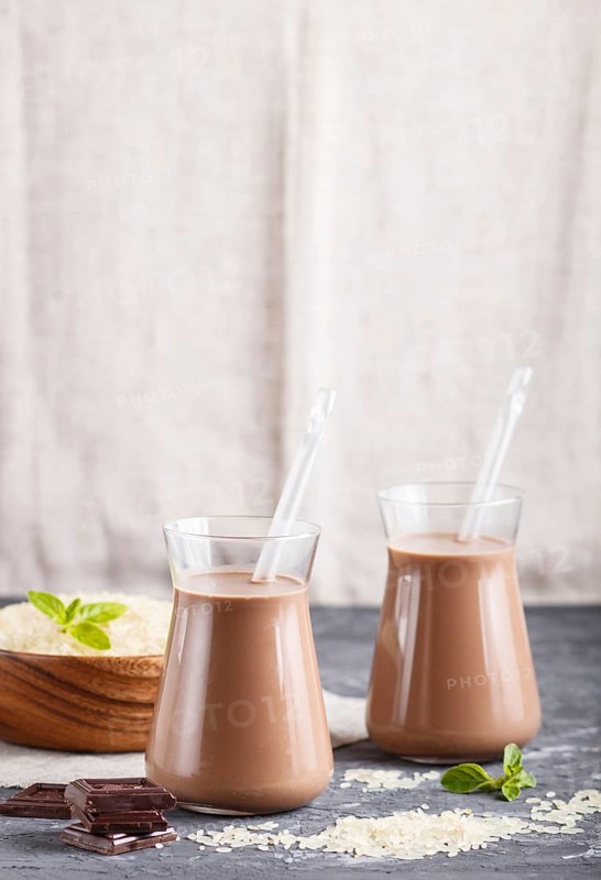 Organic non dairy rice chocolate milk in glass and wooden plate with rice seeds on a black concrete background. Vegan healthy food concept, close up, side view, copy space