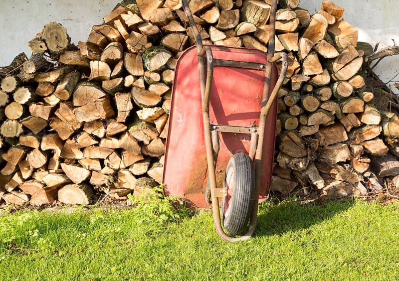 Old red metal wheelbarrow leaning against pile wood logs in garden, Cherhill, Wiltshire, England, UK