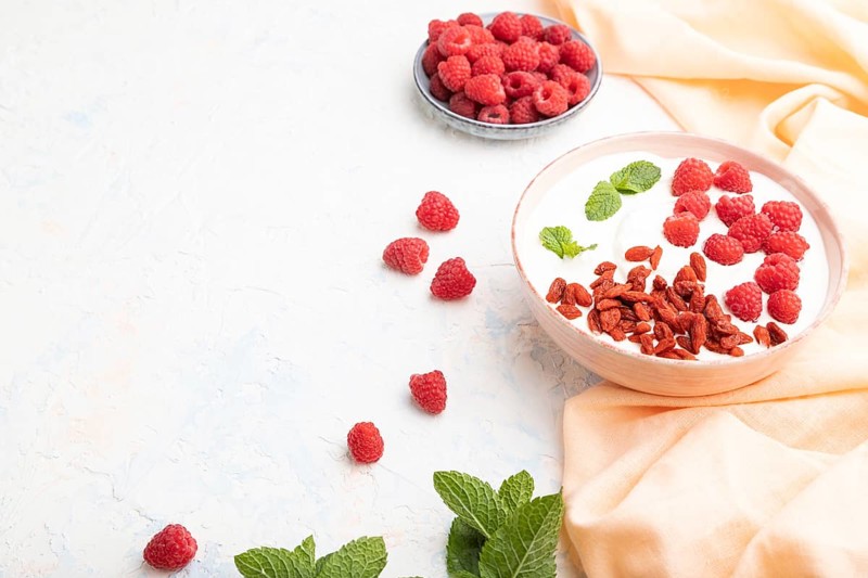 Yogurt with raspberry and goji berries in ceramic bowl on white concrete background and orange linen textile. Side view, copy space