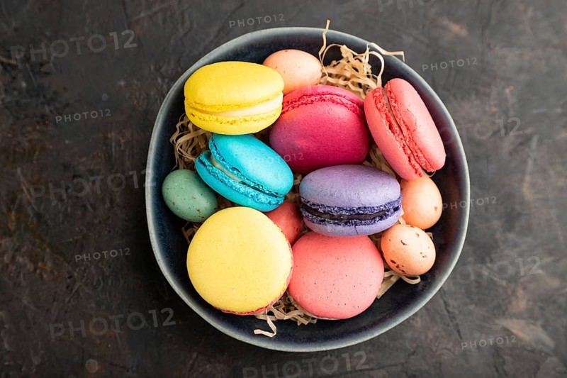 Multicolored macaroons and chocolate eggs in ceramic bowl on black concrete background. top view, flat lay, close up, still life. Breakfast, morning, concept