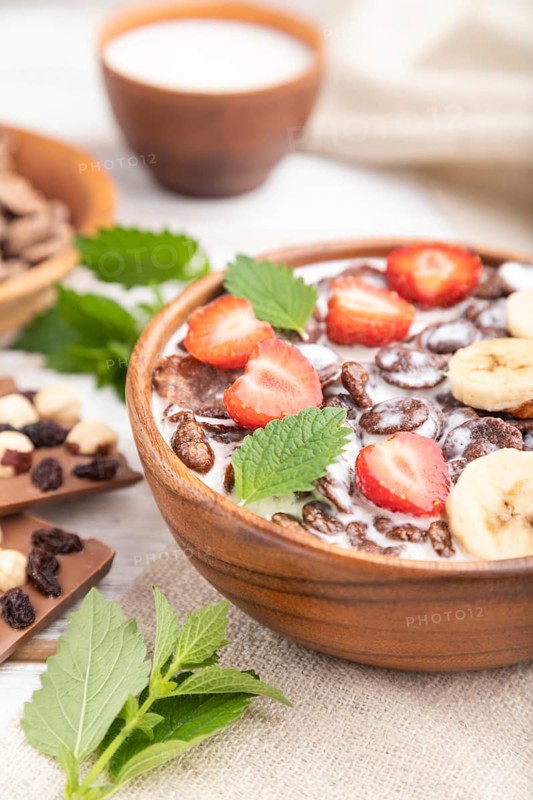 Chocolate cornflakes with milk and strawberry in wooden bowl on white wooden background and linen textile. Side view, close up, selective focus