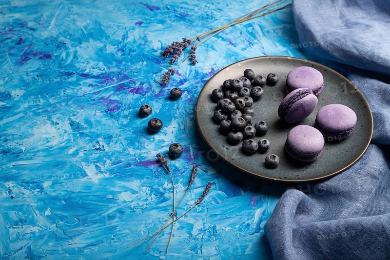 Purple macarons or macaroons cakes with blueberries on ceramic plate on a blue concrete background and blue textile. Flat lay, side view, copy space