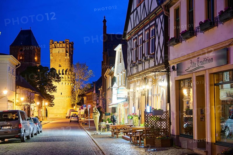The Lange Straße with half-timbered houses and other old buildings, covered with cobblestones, illuminated at night. The Neustädter Tor in the background. Old town centre of Tangermünde, Hanseatic town in the Altmark. Saxony-Anhalt, Germany, Europe