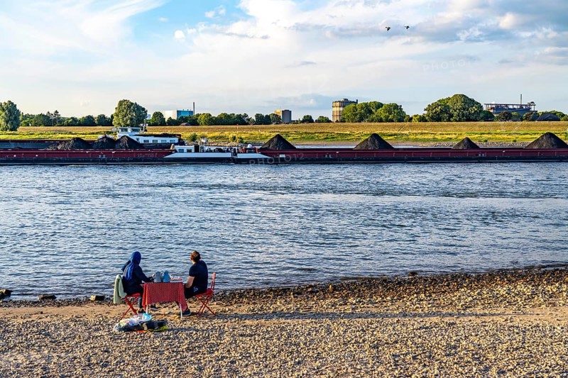 The Rhine at Duisburg-Baerl, man and woman having a picnic on the Rhine beach, at low tide, on the other side of the Rhine the Thyssenkrupp Steel steelworks and Duisburg-Beeckerwerth, cargo ships, coal freighters, Duisburg, North Rhine-Westphalia, Germany, Europe
