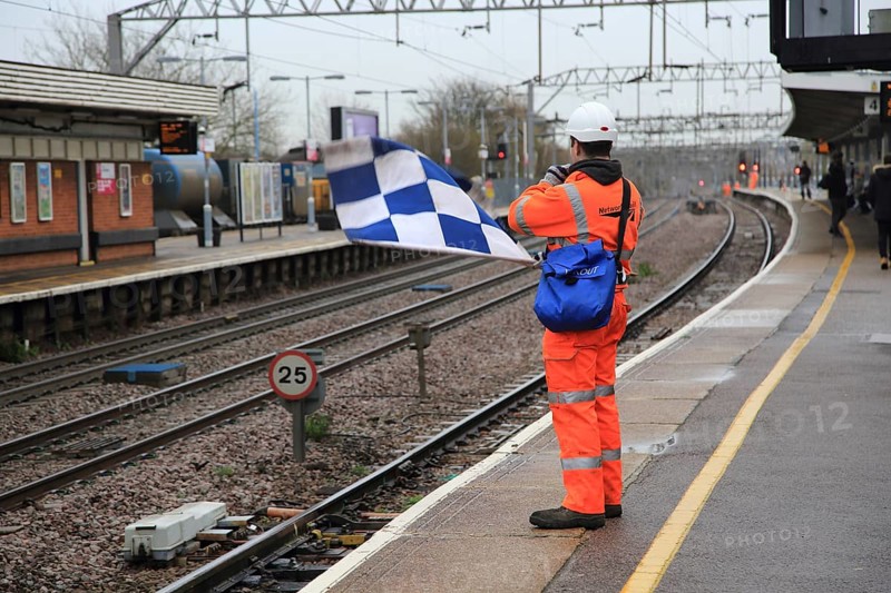 Railway worker wearing high visibility orange clothing holds blue and white checkered safety flag, Colchester, Essex, England, UK