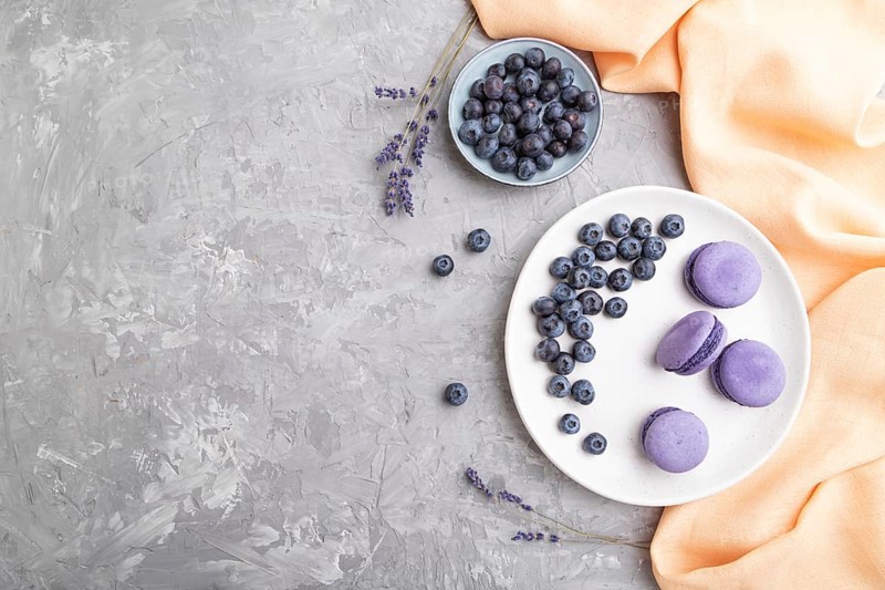 Purple macarons or macaroons cakes with blueberries on white ceramic plate on a gray concrete background and orange textile. Flat lay, top view, copy space