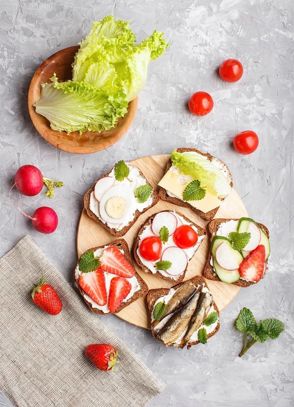 Set of different sandwiches with cheese, radish, lettuce, strawberry, sprats, tomatoes and cucumber on wooden board on a gray concrete background. top view, flat lay