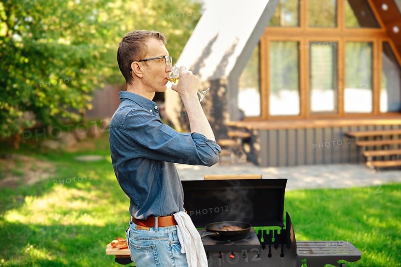 Middle-aged man drinks wine while standing in profile at the grill in the backyard of a log cabin