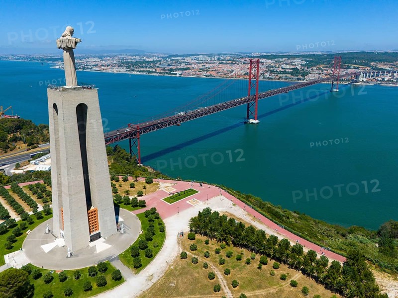 A tall statue with outstretched arms stands on a viewpoint near a bridge on the water, with a city in the background, aerial view, Statue Cristo Rei, Almada, Ponte 25 de Abril, 25 April Bridge, suspension bridge over the Tagus, double-decker bridge, road traffic and railway traffic, Lisbon, Lisboa, Portugal, Europe
