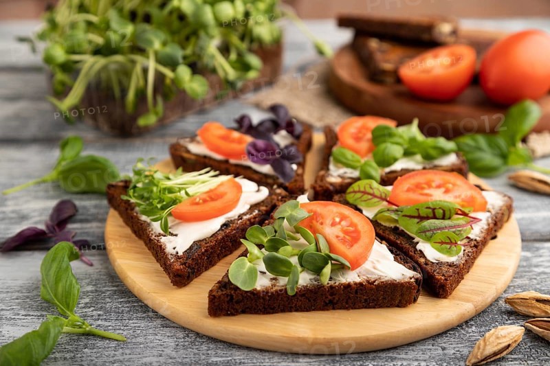 Grain rye bread sandwiches with cream cheese, tomatoes and sunflower microgreen on gray wooden background and linen textile. side view, close up, selective focus
