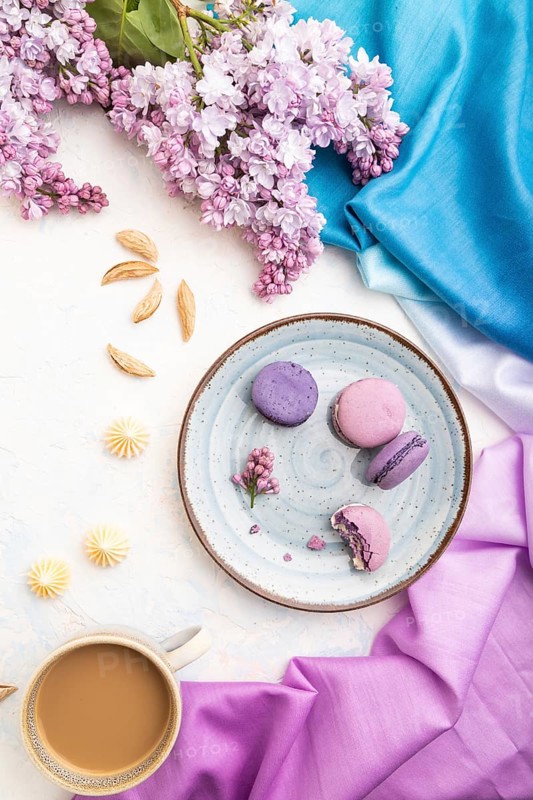 Purple macarons or macaroons cakes with cup of coffee on a white concrete background and magenta-blue textile. Top view, flat lay, close up